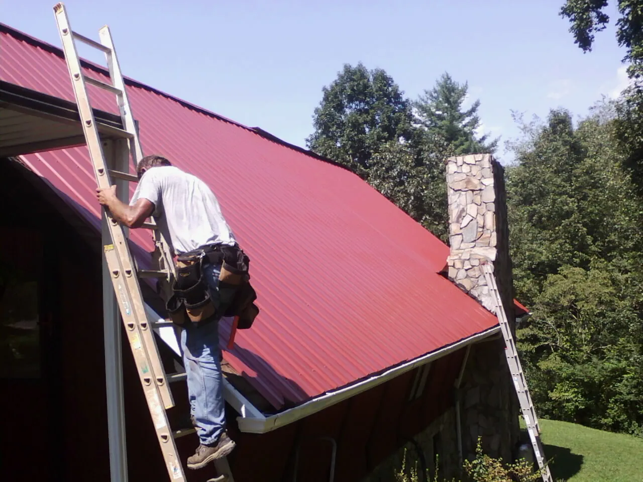 Roofer on a ladder beside a red metal roof and stone chimney — Soffit & Fascia Repair by Roofing Brothers in South Londonderry